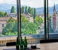 A modern room with large windows offering a scenic view of a cityscape and mountains, featuring a wooden table with three green bottles and several black chairs.