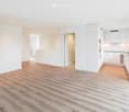 Empty room with light wooden flooring, white walls, and a modern kitchen area with white cabinets and stainless steel appliances visible in the background.