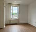 an empty room with white walls, wooden floor, and an open window showing a view of houses and greenery outside