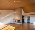 Large open room with wooden flooring, tall wooden ceiling, central brick fireplace with mantle, sliding door to the garden, white walls