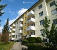 Multi-story residential building with white balconies and yellow facade, surrounded by greenery and a paved walkway.