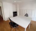 A modern, minimalist office space with a white table, black office chair, and a small black cabinet. The room features light wooden flooring and white walls with paneling.