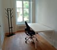 A small, well-lit room with wooden flooring, a white table, a black office chair, and a coat rack near the door.