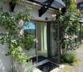 A modern house entrance with large glass doors, surrounded by lush greenery and climbing roses. The pathway is gravel, and the entrance features a doormat.