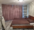 A modern bedroom with a large bed, wooden headboard, and brown bedding. Features include a guitar leaning against a white cabinet, a nightstand with a lamp, and large windows with brown curtains.