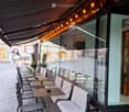 An outdoor seating area with tables and chairs, featuring a covered canopy with string lights, located on a cobblestone pavement next to a building with large windows.