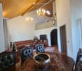 Spacious living and dining area with exposed wooden beams, chandeliers, a sofa, coffee table, dining table with chairs, and a television on a wooden stand.