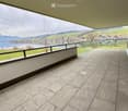 Spacious balcony with modern tiled flooring, expansive views of a lake and surrounding mountains, glass railing, and a grassy area.