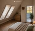Cozy attic bedroom with wooden slanted ceilings, skylights, and large windows. Features a bed with pillows and a blanket, a nightstand with a lamp, a drawer, and a potted plant near the window.