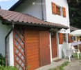 A white house with wooden shutters and a tiled roof, featuring a small patio area with white plastic chairs, a table, and an umbrella.