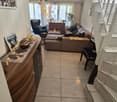 Living room with beige tiled floor, white walls, and a staircase leading upwards. Features a beige sectional sofa, coffee table, wooden cabinet, and a blue armchair.