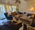 Cozy living room with a sofa, armchair, coffee table, and a small table with a laptop. The room features a large window with a view, a chandelier, and several decorative plants.