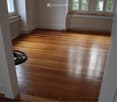 Empty room with polished wooden flooring, white walls, and radiators along the walls. Natural light streams in from windows at the far end.
