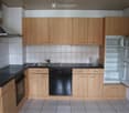 Modern kitchen with light wooden cabinets, black countertops, stainless steel appliances including a refrigerator, dishwasher, and oven, and a sink with a faucet.