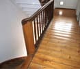 Wooden staircase with polished wooden handrail and balusters, leading up to a landing area with white walls and a closed door at the end.