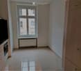 Empty room with tiled floor, light-colored walls, and a window with double doors. There is a radiator below the window and a kitchen area visible on the left side with cabinets and an oven.