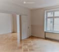 Empty room with light wooden flooring, white walls, and a large window with a radiator below it. There is a door leading to another room.
