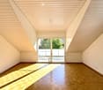 Bright, empty room with wooden flooring and slanted wooden ceiling. Large glass doors lead to an outdoor area with a tiled patio and greenery visible beyond.