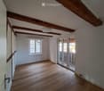 Empty room with wooden floor, exposed wooden beams on the ceiling, white walls, and a large window with white frames. A sliding glass door leads to a balcony or terrace area.
