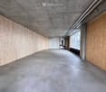 Modern, minimalist corridor with concrete floors and walls, wooden paneling on one side, and large windows allowing natural light to enter.