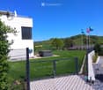 A modern house with a well-maintained lawn, a hot tub, and a metal gate. The background features rolling hills and flags on poles.