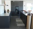 Modern kitchen with dark grey cabinets, stainless steel appliances, island with integrated stove, sink, and seating area. Features a small potted plant, knife block, and decorative columns.