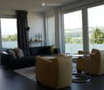 Modern living room with large windows offering a scenic view, featuring a black leather sofa, cream armchairs, a round glass coffee table, and a small side table with decorative items.