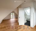An unfurnished attic room with wooden flooring, slanted ceilings, and a door leading to a small bathroom. The bathroom features a toilet, sink, and a small window with a skylight above.