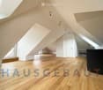 Bright, empty attic room with wooden flooring, white walls, and large windows allowing natural light. The room features a white door and built-in storage space along one wall.