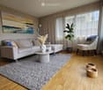 Modern living room with a grey sofa, coffee table, armchair, large windows with sheer curtains, and a potted plant. The room features a wooden floor, a grey rug, and decorative items including a picture frame and a shelf.