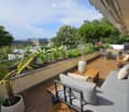 A spacious, well-maintained balcony with wooden decking, outdoor furniture including a sofa, small tables, and chairs, surrounded by lush greenery and potted plants. The view includes a cityscape with trees and buildings.