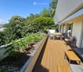 Balcony with wooden decking, outdoor furniture including a table, chairs, a sofa, and a bench, surrounded by greenery and trees.