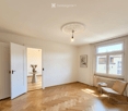 Bright, unfurnished room with wooden flooring, large windows, a single chair, a small table with a vase, and a picture frame on the wall.