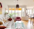 Modern living room with white walls, large windows, a white sectional sofa, a pink armchair, a glass coffee table, and a small round stool. The room features pendant lamps, a painting, and a small plant on the windowsill.