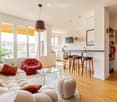 Modern, open-plan living and dining area with a white sectional sofa, red accent chair, ottoman, glass coffee table, and wooden stools at a kitchen counter. Large windows with yellow awnings provide natural light, and a small plant adds a touch of greener