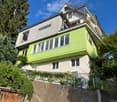 Two-story house with green and white exterior, large windows, and a balcony on the upper level.