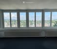 Empty room with large windows offering a scenic city view, radiators, and a dark carpeted floor.