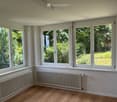 Empty room with wooden flooring, white walls, large windows overlooking a green garden, and two radiators along the wall.