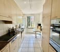 Modern kitchen with light wood cabinets, tiled floor, and stainless steel appliances. Features an island with a sink, stove, and microwave, along with a small dining area with a table and chair.