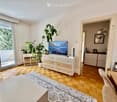 Bright living room with wooden flooring, large sliding glass doors leading to a balcony, television on a wooden cabinet, potted plants, and a small seating area near the balcony.