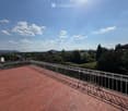 Brick-paved balcony with metal railing, overlooking a suburban area with trees and buildings.