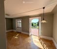 Empty room with wooden herringbone flooring, neutral-colored walls, and large windows. A hanging pendant light is visible, and an open door leads to a balcony or terrace area.
