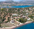 Aerial view of a coastal village with scattered houses, trees, and a lake in the background, surrounded by hills and mountains.