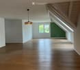 Empty attic space with wooden flooring, white walls, and a single hanging light fixture. A window provides natural light, and the sloped ceiling features exposed wooden beams.