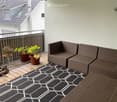 Balcony with wooden decking, black and white geometric rug, wicker sofa and chairs, small table with potted plants, and a view of neighboring building.