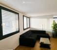 Modern living room with large windows, black sectional sofa, light-colored coffee table, and a potted plant in the corner.