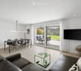 Modern living and dining area with a black leather sofa, glass coffee table, dining table with chairs, and a large TV mounted on the wall. Large sliding glass doors open to a patio with outdoor furniture.
