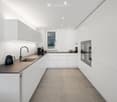 Modern, minimalist kitchen with white cabinets, dark countertops, and built-in appliances. Features a sink, faucet, coffee maker, refrigerator, kettle, and fruit bowl on the counter.