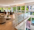 Modern, open-concept living and dining area with large windows offering a view of greenery. Features include a sofa, coffee table, dining table with chairs, desk with computers, and potted plants.
