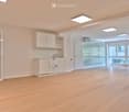 Empty room with light wooden flooring, white walls, and a small kitchenette area with cabinets and a sink. Large windows provide natural light.
